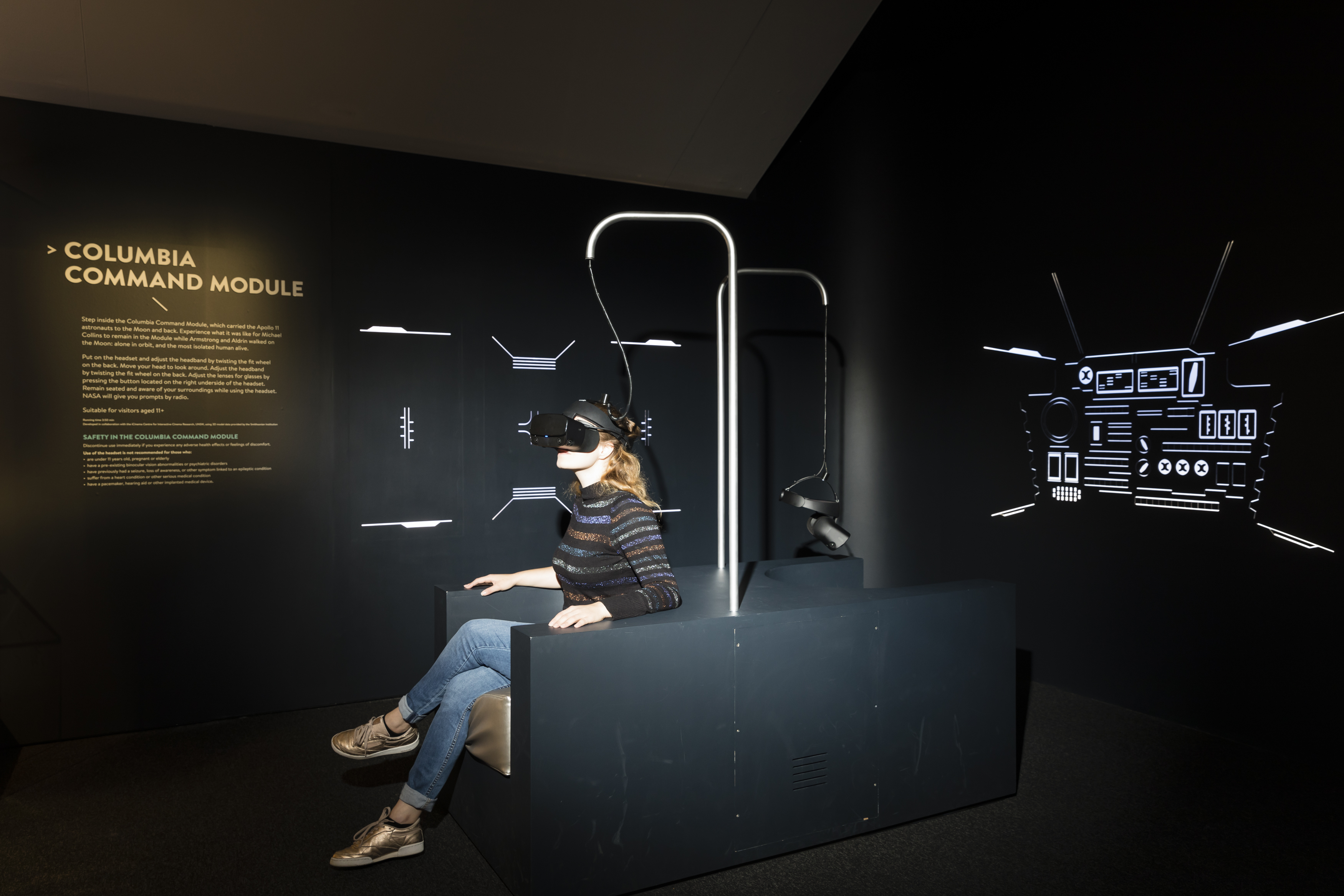 Dark nook with two seats built back-to-back. Above each seat dangles a VR headset attached to a metal pole. A woman sits in one seat wearing the headset. On the walls is an instructional text panel and backlit schematics of the Columbia control panels.